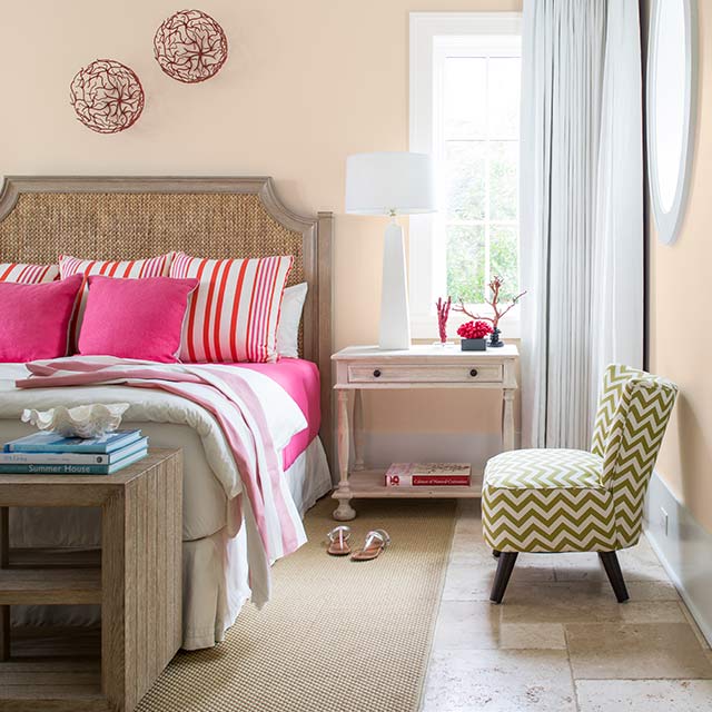 Une chambre à coucher aux murs peints en pêche arborant une chaise à chevrons verts, une table de chevet et un lit recouvert de literie rose.