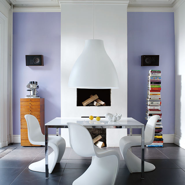 A modern dining room with a purple-painted accent wall, white fireplace, stacks of books, a dining table, and 3 modern dining chairs.