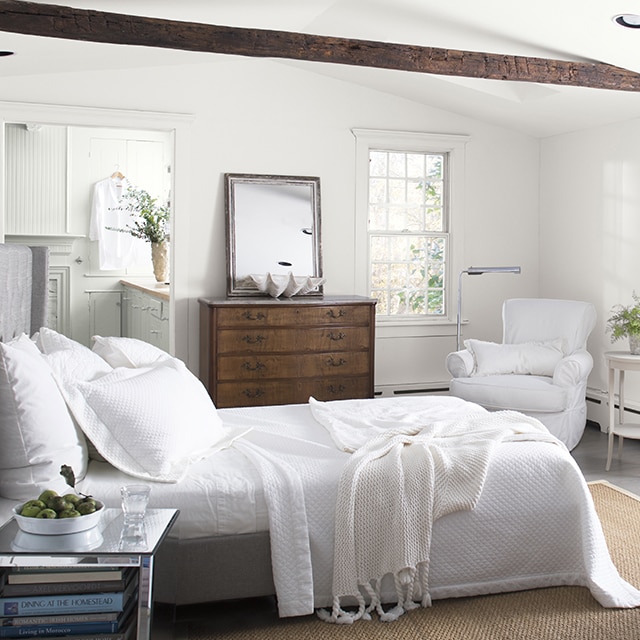 A cozy, white-painted bedroom with white bedding and chairs, a bay window with white cushions, an antique dresser, and a vaulted white ceiling with a dark brown wooden beam, looking into a white bathroom.