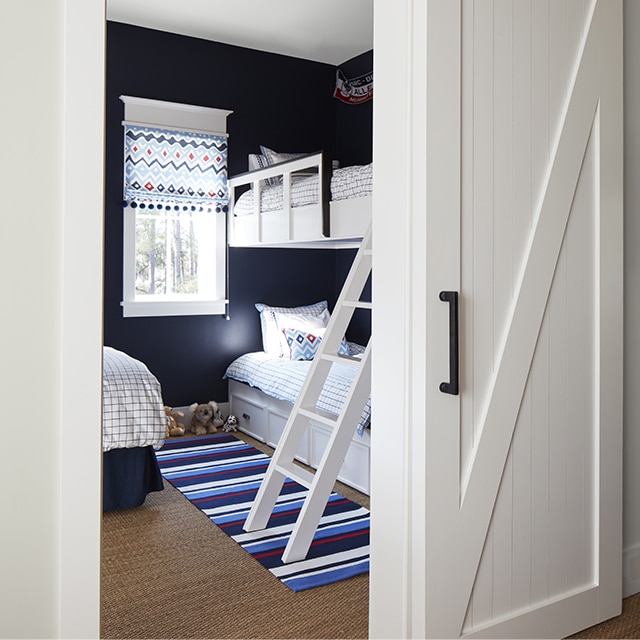 A charming kids bedroom painted in navy blue and white with a bunkbed, striped rug and sliding white barn door.