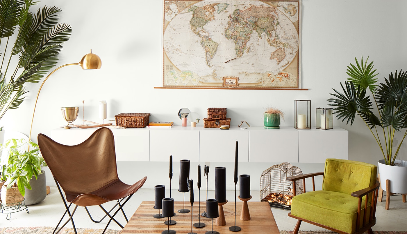 A bright loft living room painted in the same white hue using different sheens with a world map over modern wall-set cabinetry, a leather butterfly chair, a wood cocktail table with black candles, and several planters.
