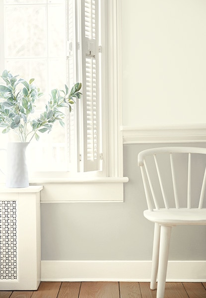 A hallway sitting area in a range of all-white hues with a two-tone painted wall, wall molding, a paned window, white shutters, a white-painted spindle-back chair and wood floor.