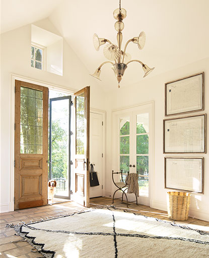 A sunlit, off-white painted entryway with vaulted ceiling, ornate chandelier, a woven beige and black area rug, and a dog looking in from one of two wooden front doors.