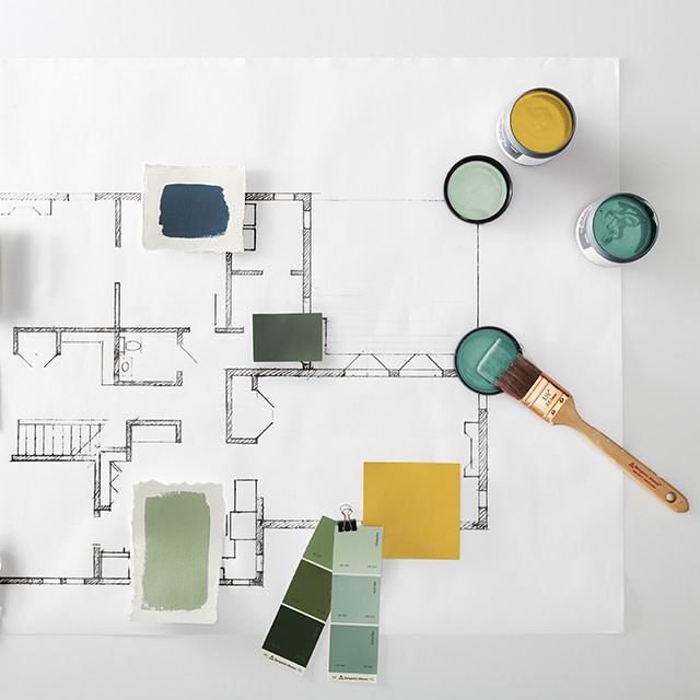 Plusieurs échantillons de couleurs Benjamin Moore, des nuanciers et des pots ouverts de peinture verte, jaune, bleue et fauve ainsi qu’un pinceau et un bâton de mélange, le tout déposé sur un plan d’étage.