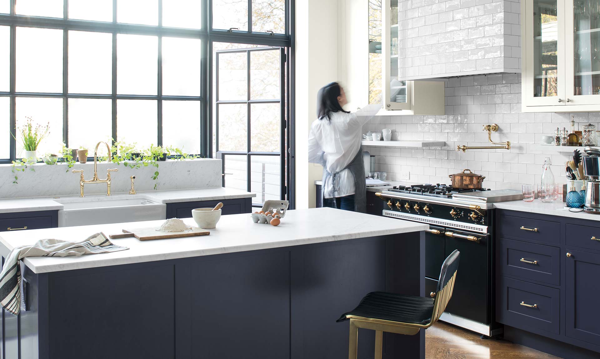 A woman reaches into a white-painted cabinet in an airy kitchen with a navy island. Colour of the Year 2019