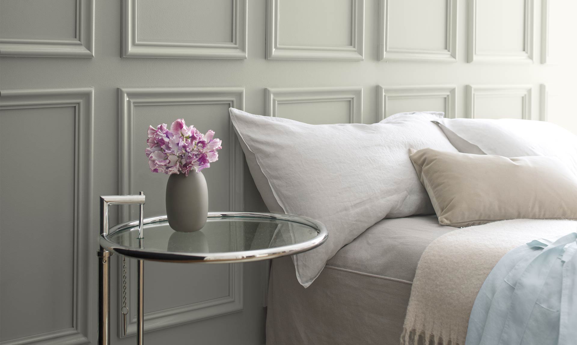 A bed against a gray wall with neutral-colored linens next to a glass side table. Color of the Year 2019