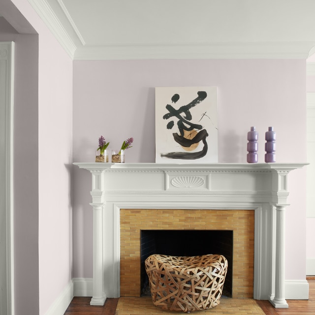 Pretty light violet-painted corner walls, white crown molding, trim and fireplace mantel with modern artwork and purple vases, and a white hallway door on the left.