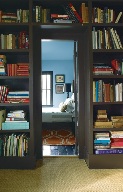 A doorway into a welcoming, blue-hued bedroom lit by natural light is framed by dark wood shelving featuring a large collection of books.