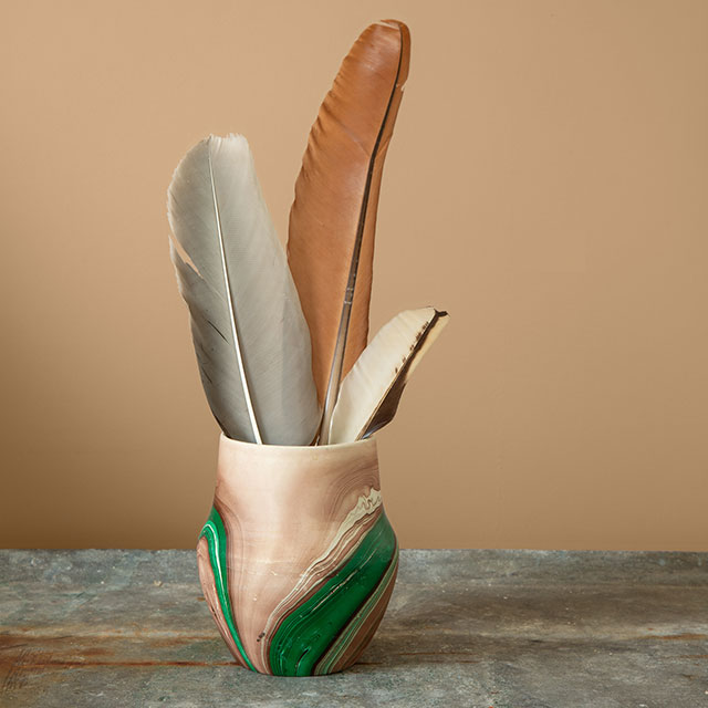 A green and cream-colored vase holding feathers on a tabletop in front of a tan painted wall.