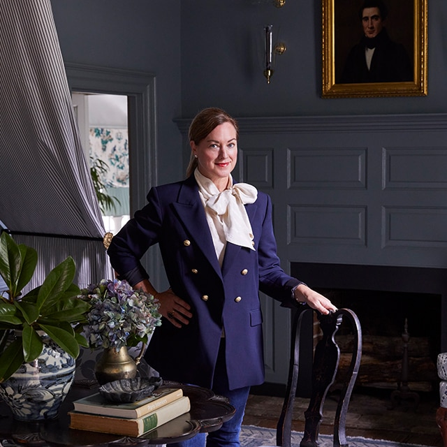 Interior designer Heather Chadduck Hillegas stands in a blue-painted study in front of a fireplace, resting a hand on the back of a traditional wooden chair next to a round, black side table.