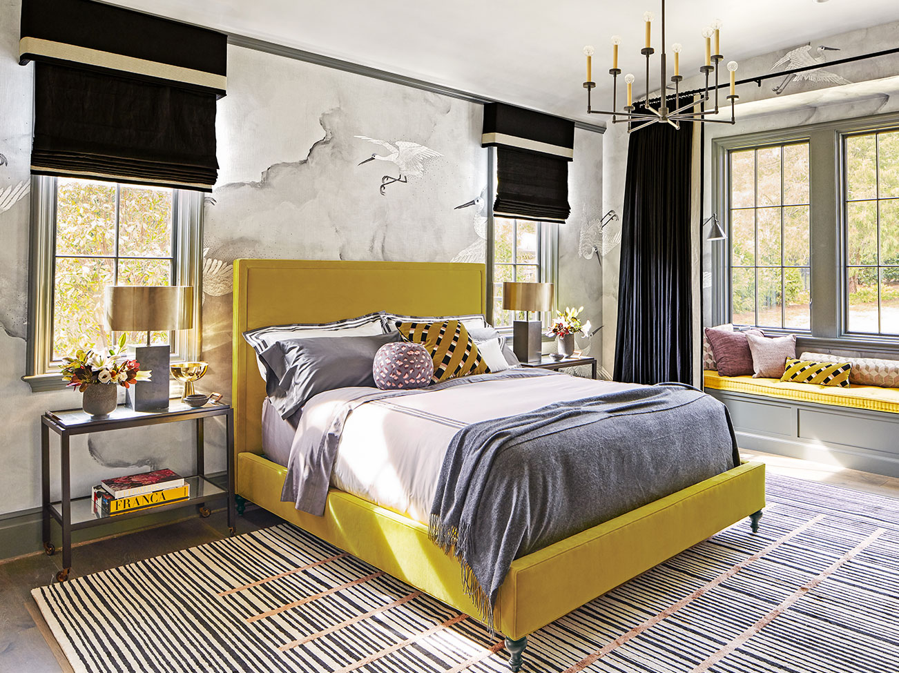 Bedroom with cloud mural, black window treatments, striped area rug, mustard yellow upholstered bed and window seat.