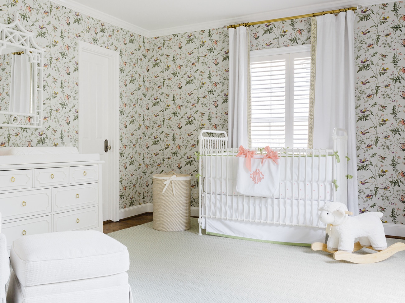 Cheery floral-papered nursery with floor-to-ceiling white drapes, white crib, dresser, chair, and white plush baby rocker.