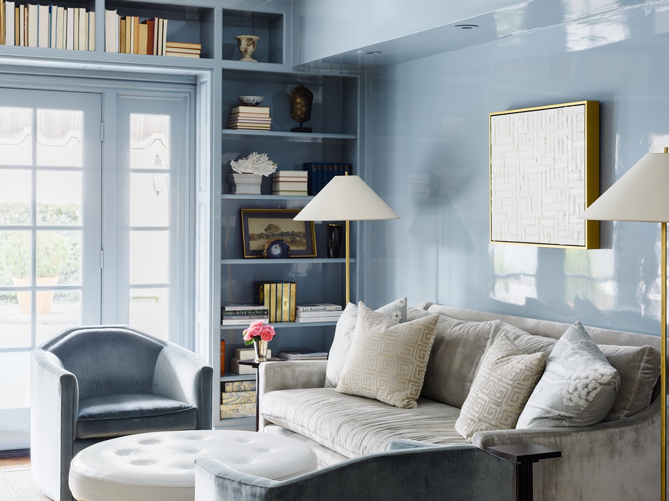 A pale blue, high gloss-painted study, gray couch, white leather ottoman, blue chairs, and a light-filled French door.