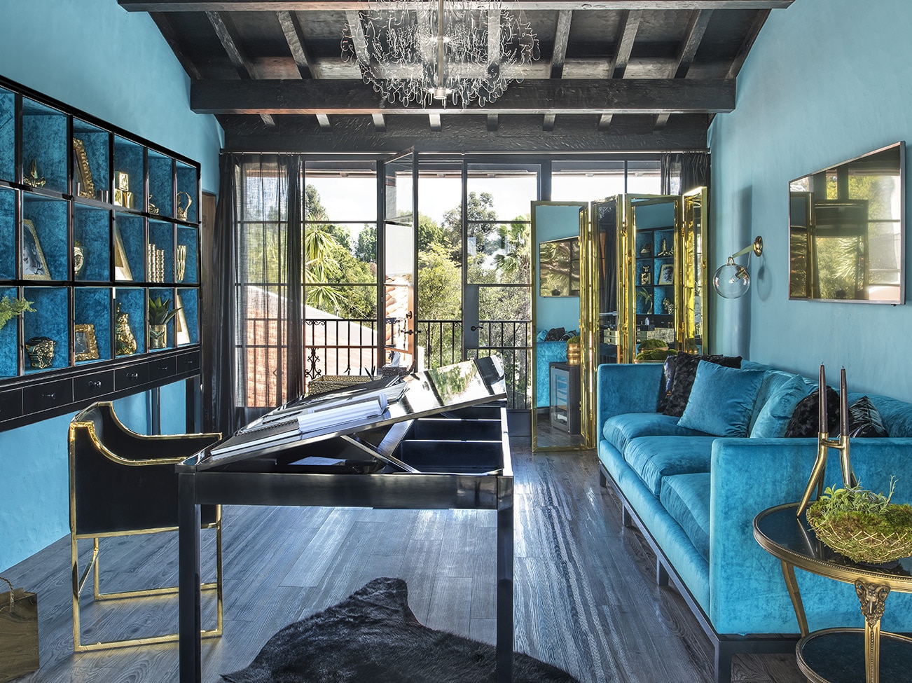 Pièce turquoise avec plafond à poutres noires, sofa tuxedo bleu, table à bascule en ébène et portes ouvrant sur une vue extérieure.