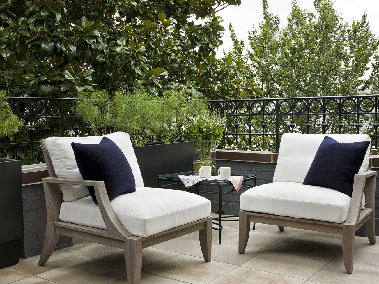 Stone patio with decorative wrought iron railing, white cushioned chairs, and navy accent pillows set against lush greenery.