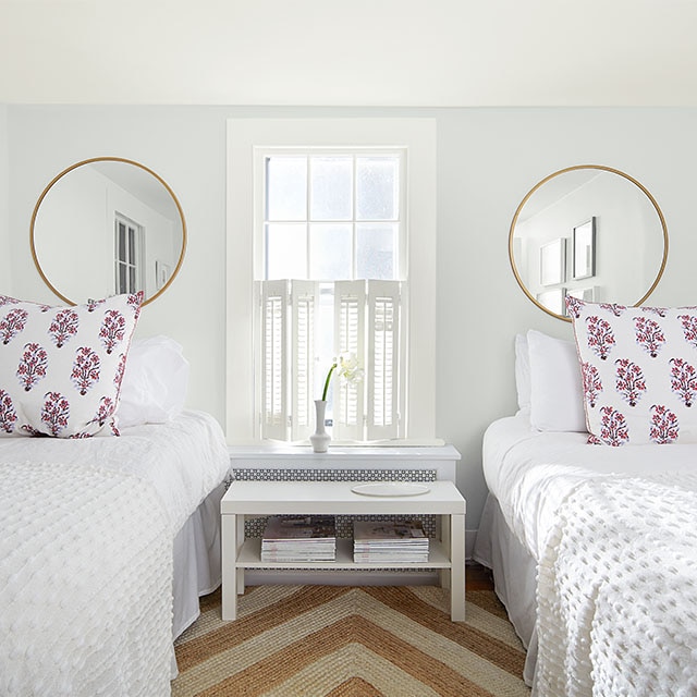 Une chambre d’adolescent blanc froid à lits jumeaux avec un coussin décoratif à fleurs roses, un miroir rond et une seule fenêtre aux moulures et volets blancs.