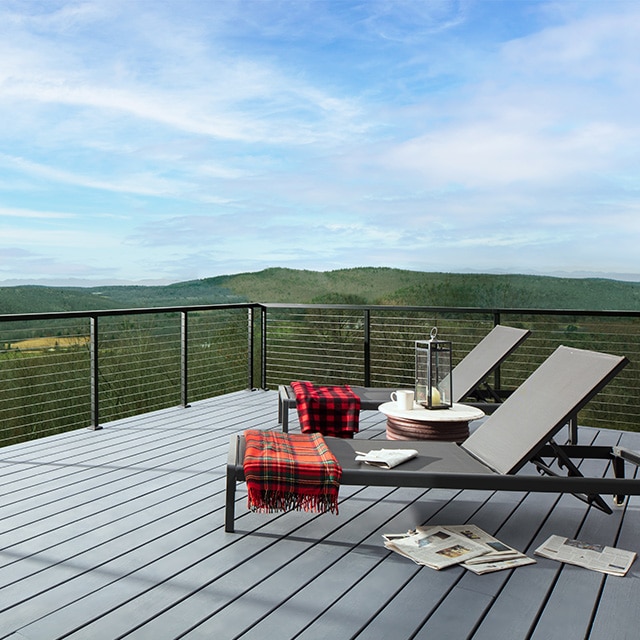 Two folding lounge chairs with draped red blankets on a light gray stained deck and a green mountainscape in the background.