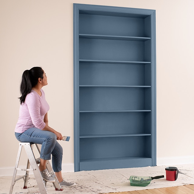 A homeowner sits on a step ladder in a pink-painted room, observing her freshly painted bookcase in a grayish blue.