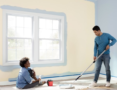 Two homeowners use a paint roller and a paintbrush to paint their beige living room blue, featuring white windows.