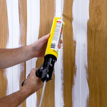 A homeowner uses caulk to fill grooves in wood paneling for a smooth finish.