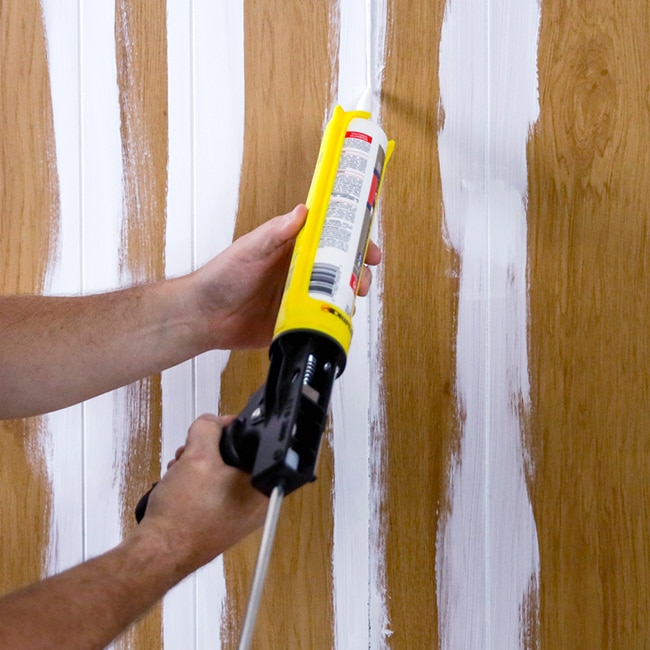 A homeowner uses caulk to fill grooves in wood paneling for a smooth finish.
