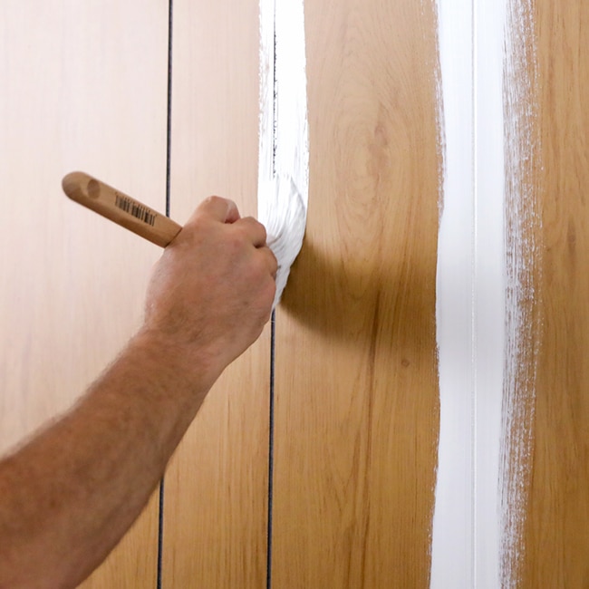 A homeowner brushes primer into the grooves of wood paneling.