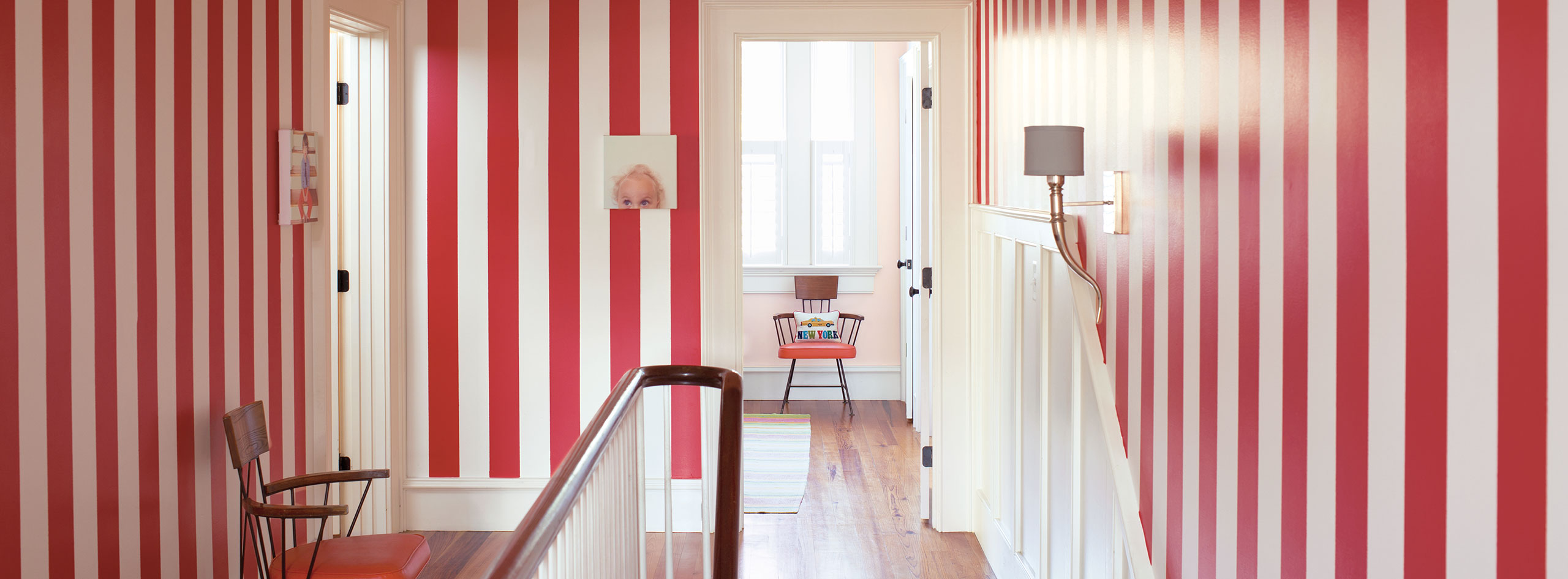 Couloir avec murs à rayures rouges et blanches, chaise des années 1950, œuvre murale originale, moulures et lambris blancs et escalier.