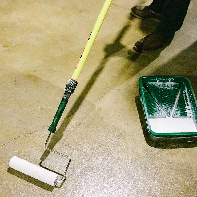 A homeowner applies sealer to a floor using a paint roller. A green painting tray is on the floor. 