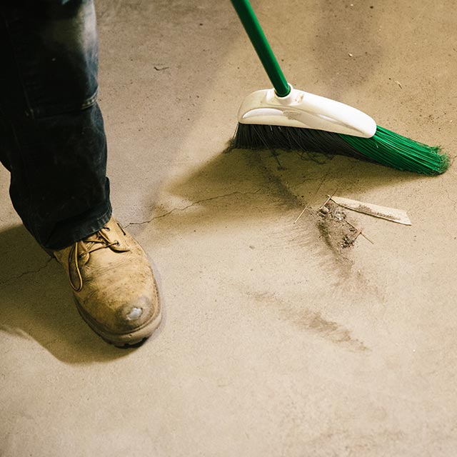 A homeowner sweeps dust and dirt from a concrete floor using a green broom.