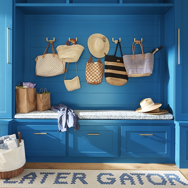 Un vestiaire peint en bleu vif avec des armoires, un banc et des tiroirs intégrés, des sacs et des chapeaux en paille suspendus et un tapis ludique bleu et blanc.