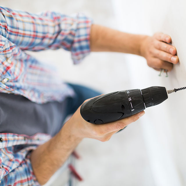 Homeowner uses a drill to insert a screw into a white-painted wall.