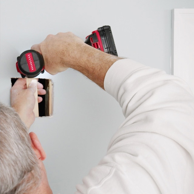 A homeowner uses a drill to secure a piece of wood to a hole in a gray wall.