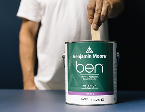 A man wearing a white shirt, stirring a can of ben Interior paint.