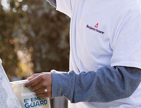 A painting contractor wearing white and blue shirts, holding a can of Element Guard™, painting the exterior of a home with a brush, and standing next to a tall ladder.