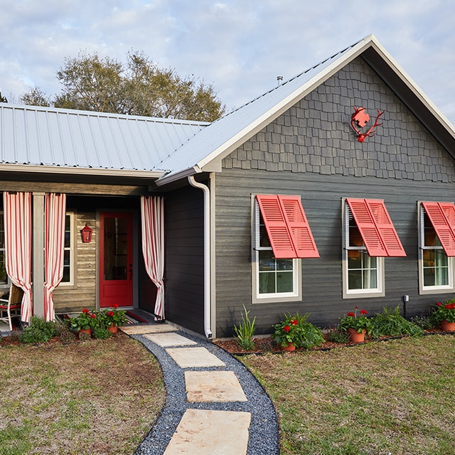 The exterior of a luxury cottage with dark, gray-painted siding, red shutters and front door, and a front porch accented with red and white striped curtains.
