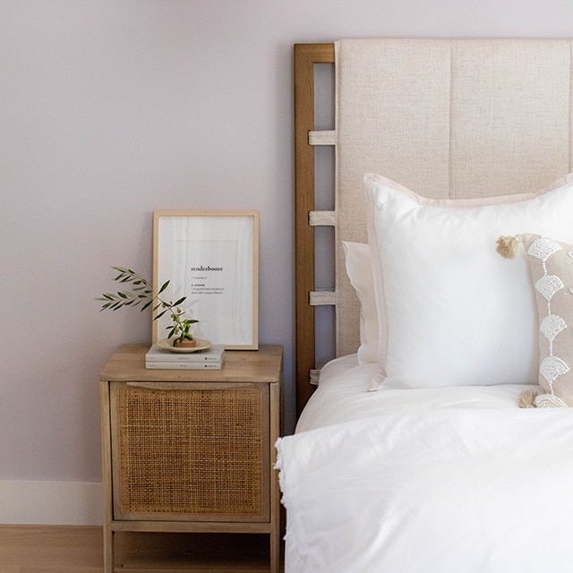 A soothing, violet painted bedroom wall with a hint of gray, a wood and beige upholstered headboard, white bedding, wood nightstand and a wicker hanging light.