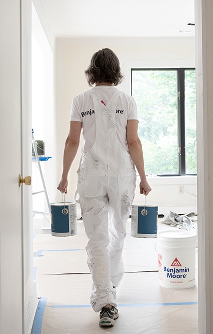 Licensed painter carrying two cans of paint walking down a hall in white overalls