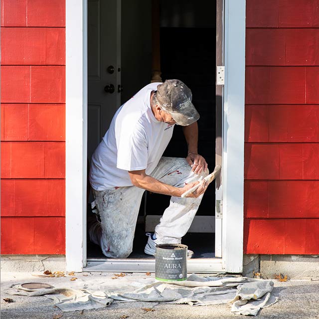 A professional painter paints the frame of a front door of a red-painted house with white Benjamin Moore paint.