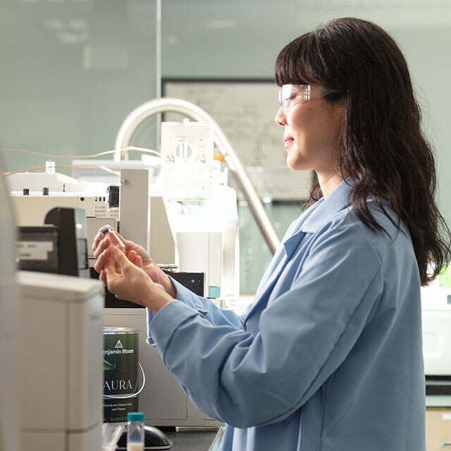 A chemist wearing safety goggles and a blue lab coat holding a glass tube, in front of lab equipment.