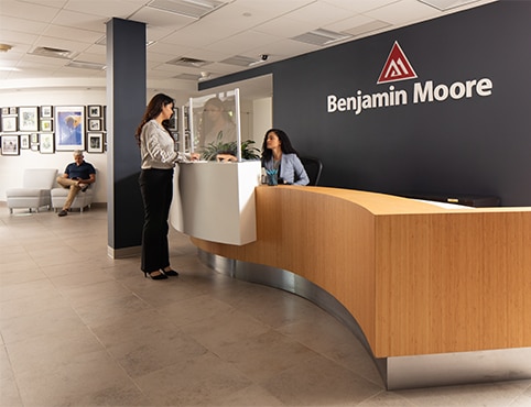 Una persona sentada detrás de un largo escritorio de recepción de madera en la oficina de Montvale de Benjamin Moore saluda a otra persona, mientras otra persona está sentada en el área de espera.