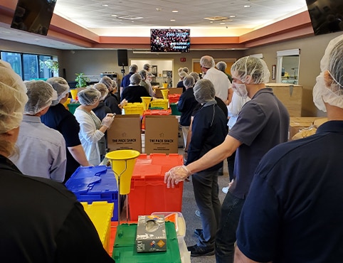Un grupo de empleados voluntarios de Benjamin Moore con contenedores de plástico de color, mirando un video instructivo sobre cómo empaquetar de forma segura kits de alimentos. Un grupo de empleados voluntarios de Benjamin Moore con contenedores de plástico de color, mirando un video instructivo sobre cómo empaquetar de forma segura kits de alimentos.