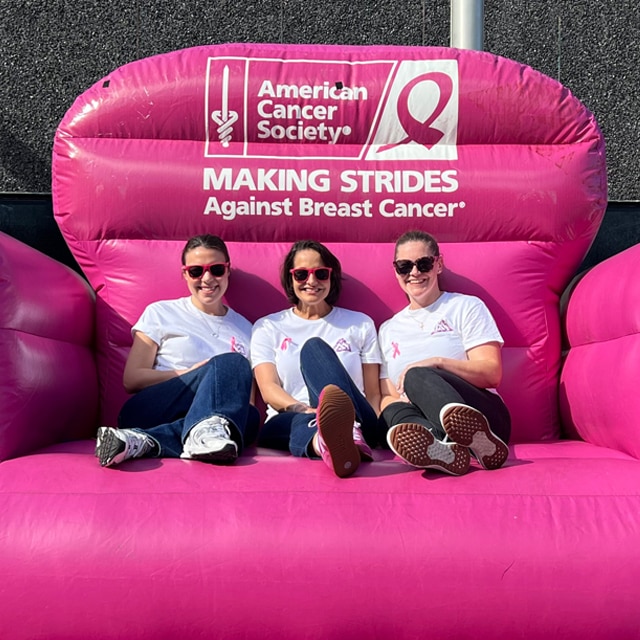 Three Benjamin Moore employees seated in a large pink inflatable chair with an American Cancer Society logo and a pink ribbon emblem.