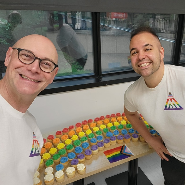 Two employees posing in front of rainbow frosted cupcakes and wearing Benjamin Moore PRISM triangle tee shirts, in celebration and support of the LGBTQIA plus community.