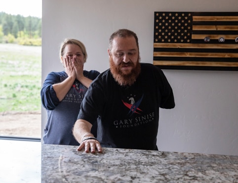 A man and woman in Gary Sinise shirts look at a granite countertop, a wooden American flag in the background.