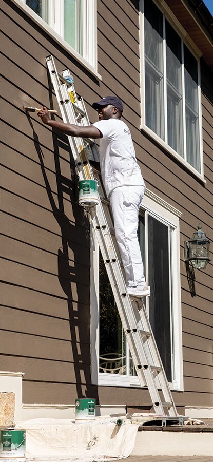A painting contractor brushes brown paint onto the exterior of a two-floor home.
