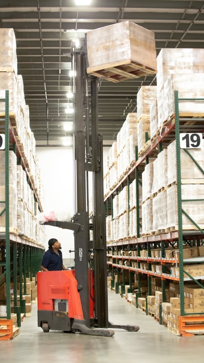 A warehouse employee driving a motorized palette machine through a warehouse. A warehouse employee driving a motorized palette machine through a warehouse.