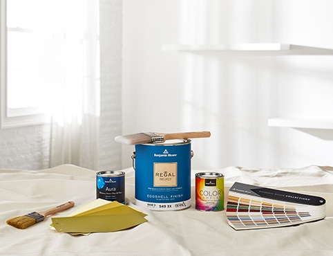 A selection of Benjamin Moore products on a drop cloth, including a gallon of Regal Select, a variety of color samples, two paintbrushes, and a fandeck.
