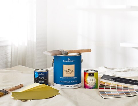 A selection of Benjamin Moore products on a drop cloth, including a gallon of Regal® Select, a variety of color samples, two paintbrushes, and a fandeck.