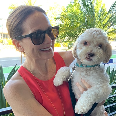 A Caucasian woman with brown hair pulled back wearing sunglasses and a red tank top holding a puppy. 