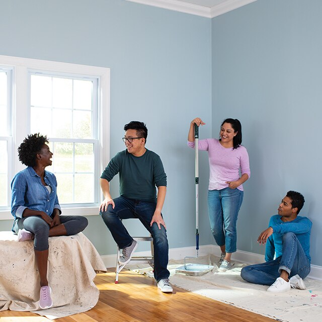 A blue-painted room with white window trim, two people sitting on drop cloths, a woman leaning with a paint roller and a man sitting on a small step ladder.